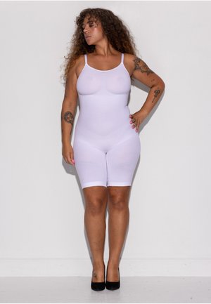 Sieviete ar cirtainiem matiem un tetovējumiem pozē, valkājot baltu pieguļošu bodiju un melnas augstpapēžu kurpes uz vienkārša balta fona.