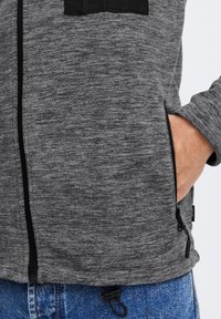 Grå zip-up jakke med en tekstureret overflade, sorte detaljer på brystlommen og justerbar snøre i kanten.