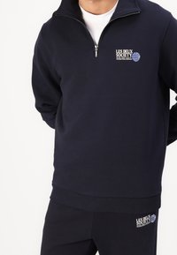 Sudadera con cremallera en color marino, hecha de una tela suave, con dobladillo y puños acanalados, y con el logo bordado de "Les Deux Society" en el pecho.