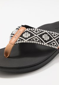 Zwarte flip-flop sandaal met een gevlochten band met een wit geometrisch patroon en een tan leren accent op de teenpost. Textuurzool.
