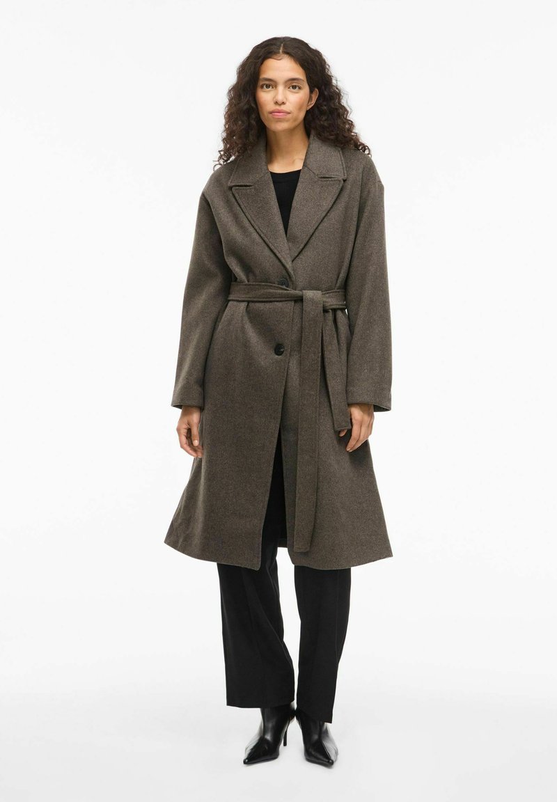 Ruskea villasekoiteviitta, jossa on leveät kaulukset, yksittäinen napitus ja vyö vyötäröllä. Yhdistetty mustiin housuihin ja korkokengiin.