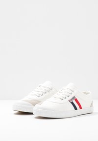 White canvas sneakers with round toe, textured rubber sole, beige accents, and red and navy striped detail on the side.