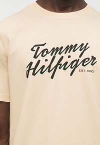 Μπεζ μπλούζα με μαύρη και τιρκουάζ σκιασμένη επιγραφή «Tommy Hilfiger» και την ένδειξη «EST. 1985» τυπωμένα στο στήθος.