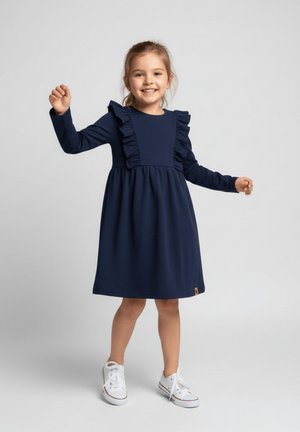 Granatowa sukienka z długimi rękawami, z falbanami na ramionach, dopasowanym gorsetem i rozkloszowanym dołem. Białe sneakersy dopełniają strój.