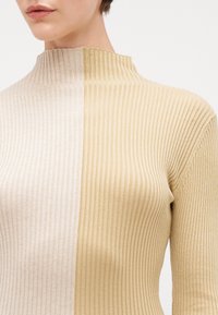 Top tricotat cu un design împărțit în bej deschis și auriu pal, cu caneluri verticale și guler înalt. Textură moale și formă ajustată.