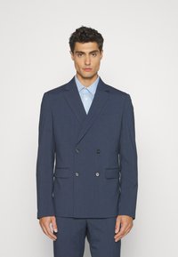 Marineblaue, doppelt geknöpfte Blazer aus glattem Stoff, mit zwei Fronttaschen, schwarzen Knöpfen und einem Reverskragen, getragen über einem hellblauen Hemd.