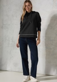 Zwarte hoodie met een voorzak en witte stiksels, gecombineerd met donkerblauwe rechttoe-rechtaan jeans en witte sneakers.