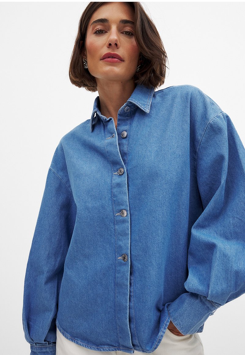 Light blue denim shirt with a button-up front, pointed collar, and long, oversized sleeves. Features metallic buttons and a relaxed fit.