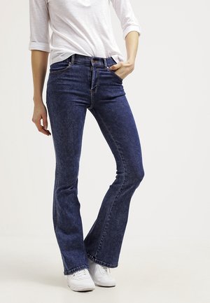 Kvinde iført mørkeblå flared jeans, hvid skjorte med op-rullede ærmer og hvide sneakers, stående med den ene hånd i lommen.