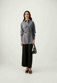 Ženska stoji v sivi kosmati srajci z pasom, črnih širokih hlačah, črnih čevljih s peto in drži majhno črno torbico.