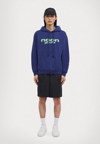 Navyblauer Kapuzenpullover mit grünem "noon goons" Text, Kordelzügen und Kängurutasche; kombiniert mit schwarzen Cargo-Shorts und weißen Sneakers.