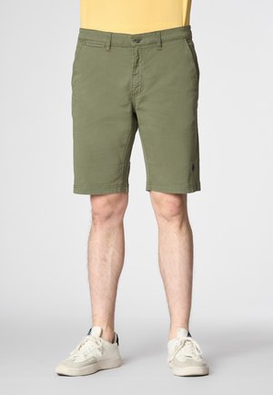 Pantaloni corti in cotone verde oliva con zip, tasche frontali e un piccolo logo all'orlo. Abbinati a scarpe da ginnastica bianche.