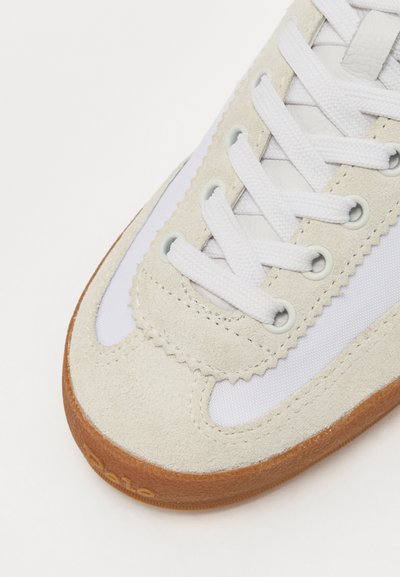 Sneaker de camurça creme com detalhes em tecido branco, apresentando atacadores redondos, costura ondulada e uma sola de borracha castanha com logotipo.