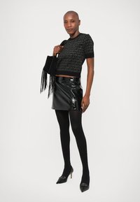  Lange vrouw in een zwart gemusterd shirt met korte mouwen, glanzende zwarte minirok, zwarte panty's, hakken en met een zwarte handtas met franjes.