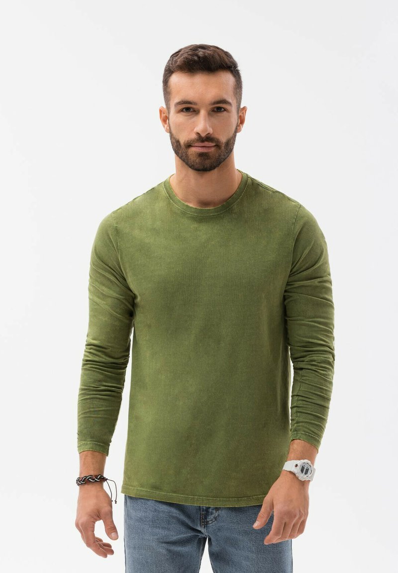 Olijfgroene langwerpige shirt met lange mouwen van een zachte, gestructureerde stof, voorzien van een ronde halslijn en aansluitende mouwen, gecombineerd met lichtblauwe spijkerbroek.