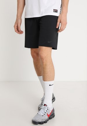 Mężczyzna ubrany w czarne spodenki Nike, białe skarpety Nike do kostek, szaro-czerwone buty do biegania Nike oraz białą koszulkę na jednolitym tle.