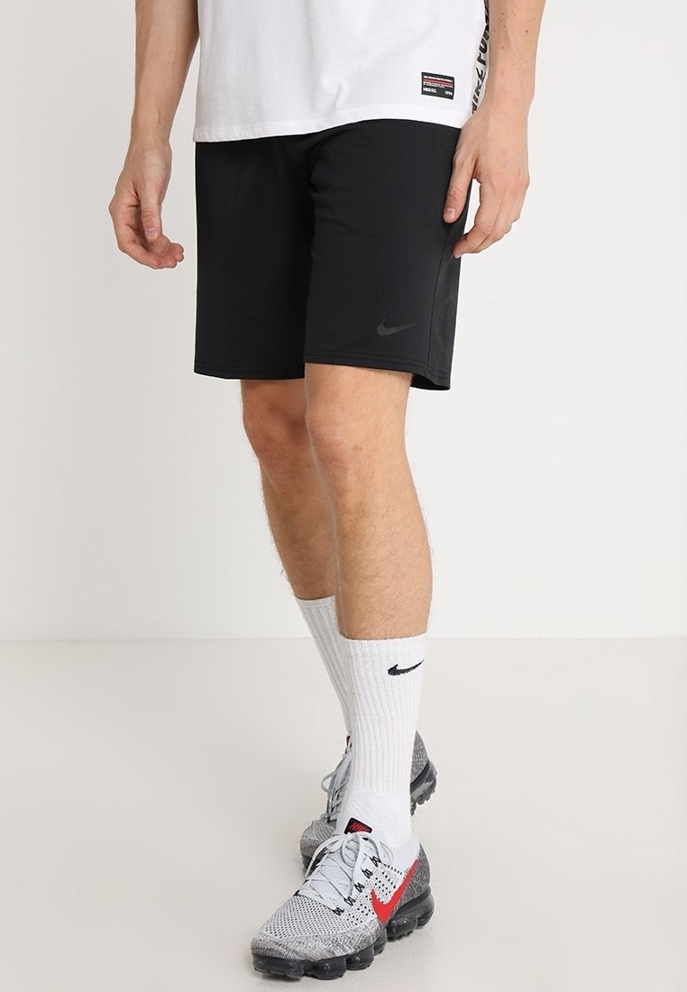 Homme portant un short noir Nike, des chaussettes montantes blanches Nike, des chaussures de course Nike grises et rouges, et un t-shirt blanc sur un fond uni.