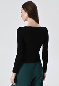 Top lungo a maniche ribattute nere con scollatura quadrata, design aderente e tessuto testurizzato. Indossato con pantaloni turchesi, visione posteriore.