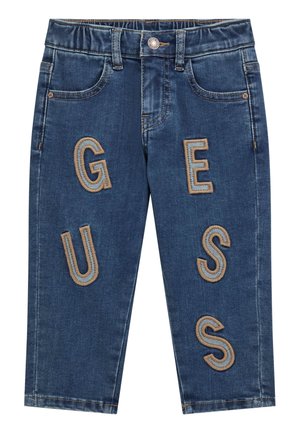 Vaqueros de mezclilla con un lavado azul oscuro, que presentan grandes letras bordadas "G," "U," "E," "S," "S" en un color tostado de contraste. Diseño clásico de cinco bolsillos.