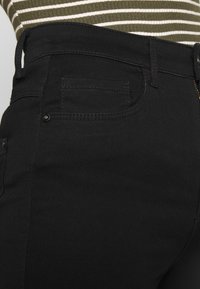 Mustat farkut, joissa on sileä rakenne, klassinen viiden taskun muotoilu ja vyölenkit vyötäröllä, sekä vetoketjukiinnitys ja nappikiinnitys.