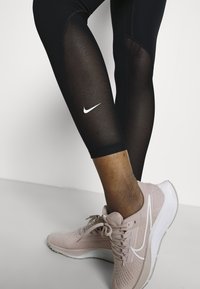 Mustat leggingsit, joissa on läpinäkyvät sivupaneelit ja valkoinen Nike-logo, yhdistettynä vaaleanpunaiseen urheilukenkään, jossa on valkoisia yksityiskohtia ja teksturoitu pinta.