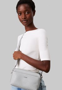 Borsa a tracolla grigia chiara con finitura testurizzata, hardware argentato e tracolla regolabile, dotata di una tasca anteriore e design elegante.