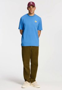 Blauw katoenen T-shirt met een geel bloemenpatroon, gecombineerd met olijfgroene ribfluwelen broek. Het model draagt een bordeauxrode pet en lichtgekleurde sneakers.