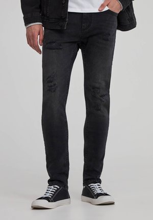 Jeans neri strappati in un taglio slim, con strappi e orli sfilacciati. Abbinati a sneakers nere e una maglietta bianca che sbuca da una giacca nera.