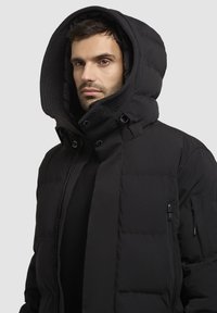 Jachetă neagră pufoasă cu guler înalt și glugă supradimensionată. Prezintă închideri cu fermoar și capse, buzunare laterale și o textură elegantă.