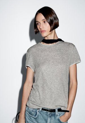 Grijze T-shirt met korte mouwen en een ronde hals, gecombineerd met lichte spijkerbroek. De stof lijkt zacht, met een subtiele textuur en naden.