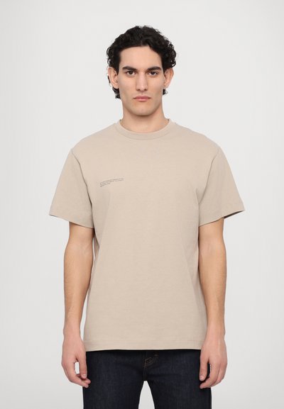 Tricou din bumbac bej, cu mânecă scurtă, guler rotund și un mic text imprimat pe partea stângă a frontalului. Purtați-l cu blugi de culoare albastră închisă.