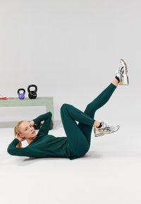 Grünes Sportoberteil und Leggings mit flachen Nähten, kombiniert mit metallischen Laufschuhen. Kettlebells und ein Springseil auf einem hellen Tisch.