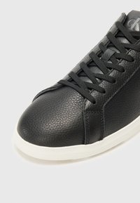 Zwarte leren sneaker met een textuur bovenwerk, zwarte veters en een witte rubberen zool met subtiele ribbels langs de rand.