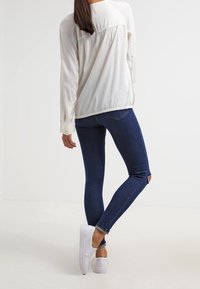 White long-sleeve blouse and dark blue skinny jeans with distressed knees. White sneakers on feet; figure stands with back turned.