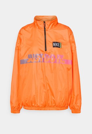Chaqueta Nike de pulóver naranja brillante con media cremallera, parche negro con el logo en el pecho y "JUST DO IT." estampado en la parte delantera en púrpura y rosa.