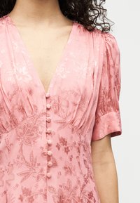 Γυναίκα που φοράει ροζ φόρεμα με μοτίβο λουλουδιών, κουμπιά καλυμμένα με ύφασμα κατά μήκος του μπροστινού μέρους και κοντά φουσκωτά μανίκια.