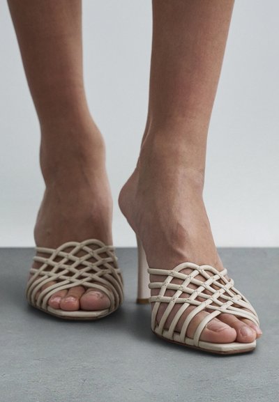 Pies con sandalias de tacón alto beige con tiras y diseño trenzado, parados sobre una superficie gris lisa contra un fondo claro.