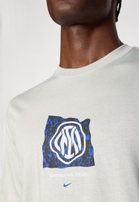 Ανοιχτό γκρι βαμβακερό t-shirt με μπλε και λευκή γραφική παράσταση του λογότυπου της Internazionale Milano. Στρογγυλή λαιμόκοψη, κοντά μανίκια, λεία υφή.