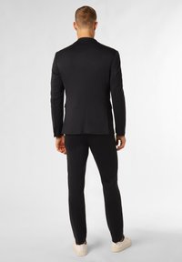 Schwarzer, maßgeschneiderter Anzug mit einer strukturierten Jacke und schmal geschnittenen Hosen aus glattem Stoff, kombiniert mit weißen Turnschuhen.