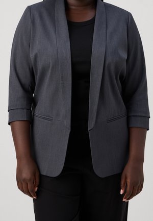 Tummanharmaa kolmen neljänneksen hihoilla varustettu bleiseri, joka on puettu mustan topin päälle henkilöllä, joka seisoo yksiväristä vaaleaa taustaa vasten.