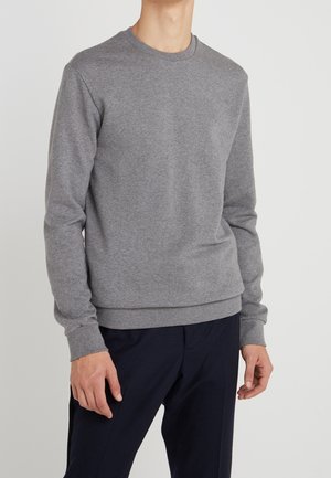 Homme portant un pull gris à manches longues et col rond, ainsi qu'un pantalon foncé, debout devant un fond clair uni.