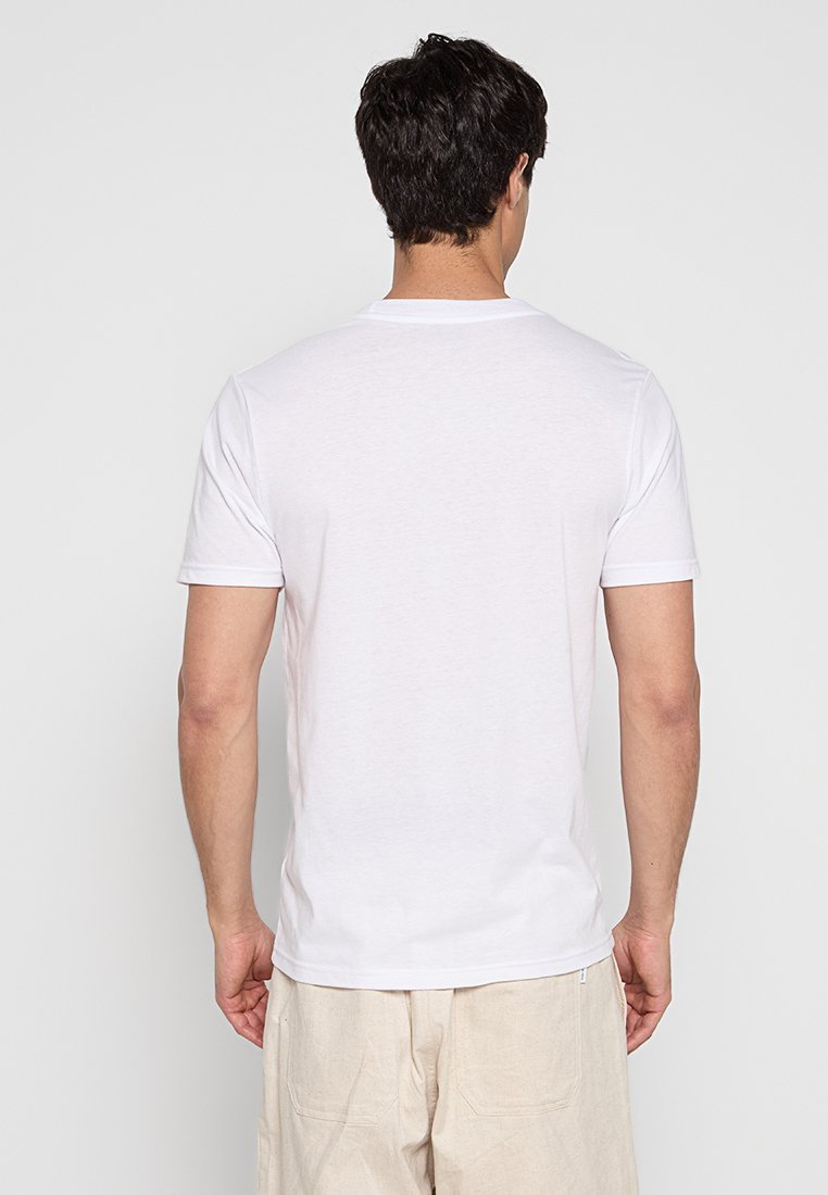 Mężczyzna z ciemnymi włosami, ubrany w gładką białą koszulkę z krótkim rękawem i beżowe spodnie, pokazany od tyłu na białym tle.