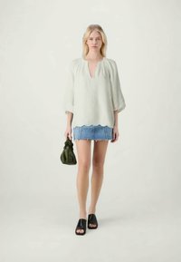 Lichtgroene blouse met een V-hals en driekwartmouwen, gecombineerd met een denim minirok en zwarte slippers, terwijl ze een groene tas vasthoudt.