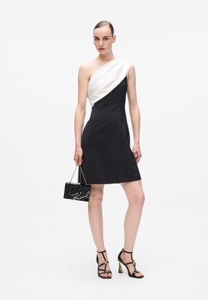 Μαύρο φόρεμα ενός ώμου με λευκή σατέν δάπεδο; λείο ύφασμα, εφαρμοστή σιλουέτα. Το μοντέλο κρατά μια μικρή μαύρη τσάντα χειρός με ασημί πούλιες και αλυσίδα.