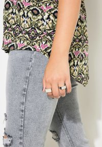 Patroon-top met groene, roze en witte bloemenmotief; lichtgrijze distressed jeans; zilveren en zwarte ringen om de hand, gladde textuur.