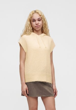 Jeune femme aux longs cheveux bouclés blonds, portant un sweat à capuche en maille beige sans manches et une jupe courte marron sur un fond uni.