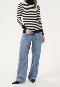 Femme portant un pull à col roulé rayé noir et blanc, un jean large déchiré bleu, et des chaussures noires à bouts pointus, debout avec la main sur la hanche.