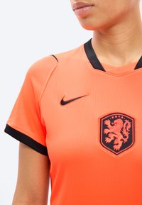 Άτομο που φοράει πορτοκαλί αθλητική φανέλα με μαύρο περίγραμμα, λογότυπο της Nike και το έμβλημα με το λιοντάρι της KNVB στο στήθος, μπροστά από ένα απλό φωτεινό φόντο.