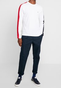 Bílý dlouhý rukáv T-shirt s červenými a tmavě modrými bočními pruhy, kulatý výstřih, nošený s tmavě modrými kalhotami a tmav�ými teniskami. Volný střih a hladká textura.