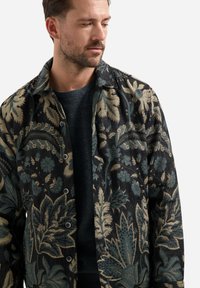 Man in een donker shirt met een groot groen en beige bloemmotief, die iets naar beneden en opzij kijkt tegen een witte achtergrond.